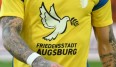 Die Welt ist entsetzt vom gewaltsamen Überfall der Ukraine durch Wladimir Putins russische Streitkräfte. Die Sportwelt bekundet ihre Solidarität mit den Opfern der Aggression - so wie hier der FC Augsburg mit einem eigens gestalteten Warmup-Shirt.
